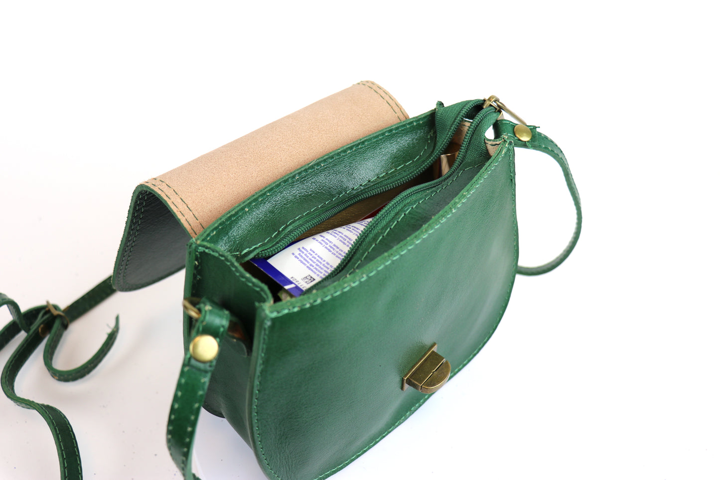 Close-up of the Cora Vintage Small Leather Crossbody Bag in green, showcasing its open interior with a visible zipper pocket. The bag is partially filled with items, including a box, and highlights the contrasting beige suede lining and brass hardware.