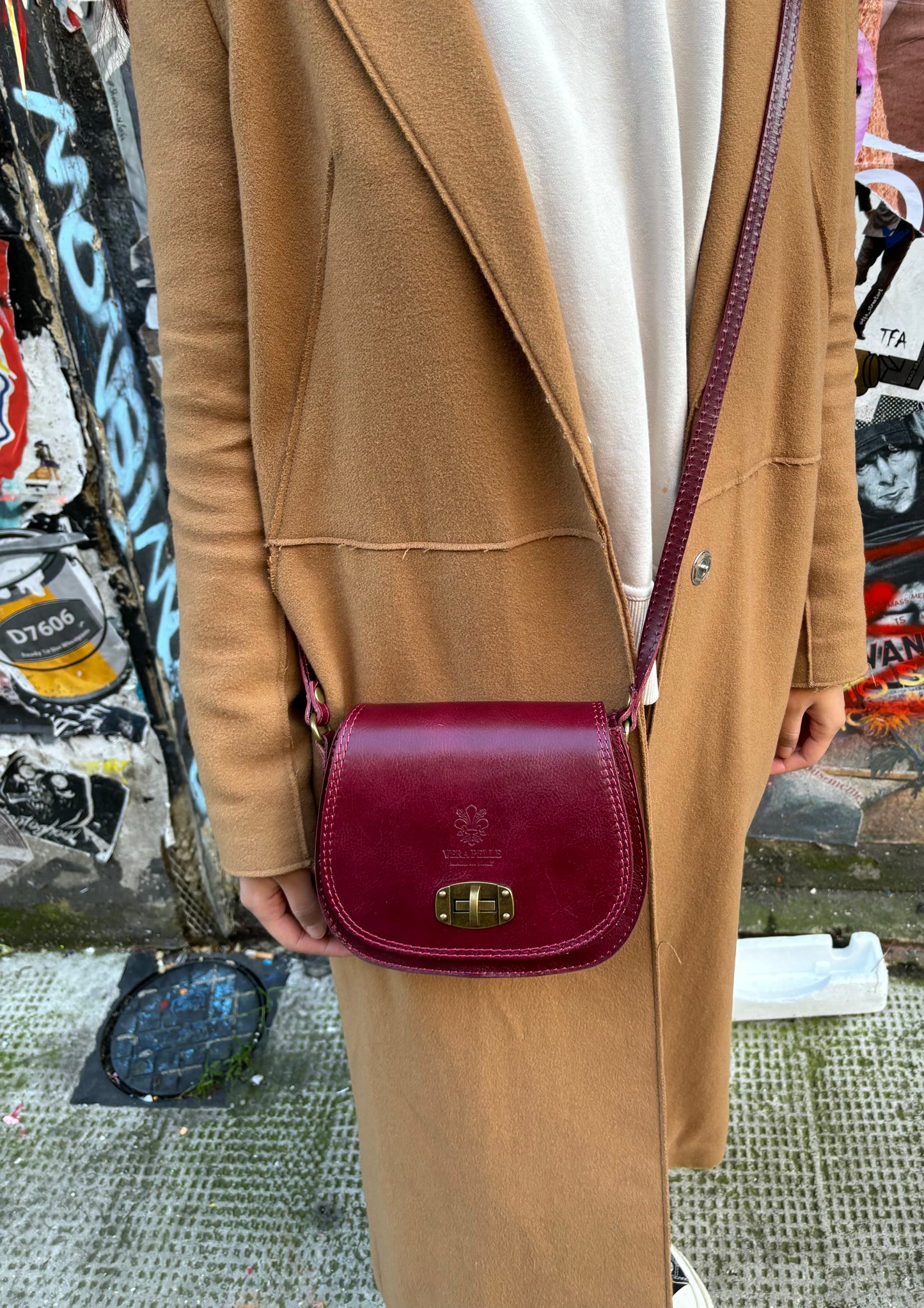 A model wearing a dark red Cora Vintage Small Leather Crossbody Bag, styled outdoors with a beige coat, against a street-art backdrop.