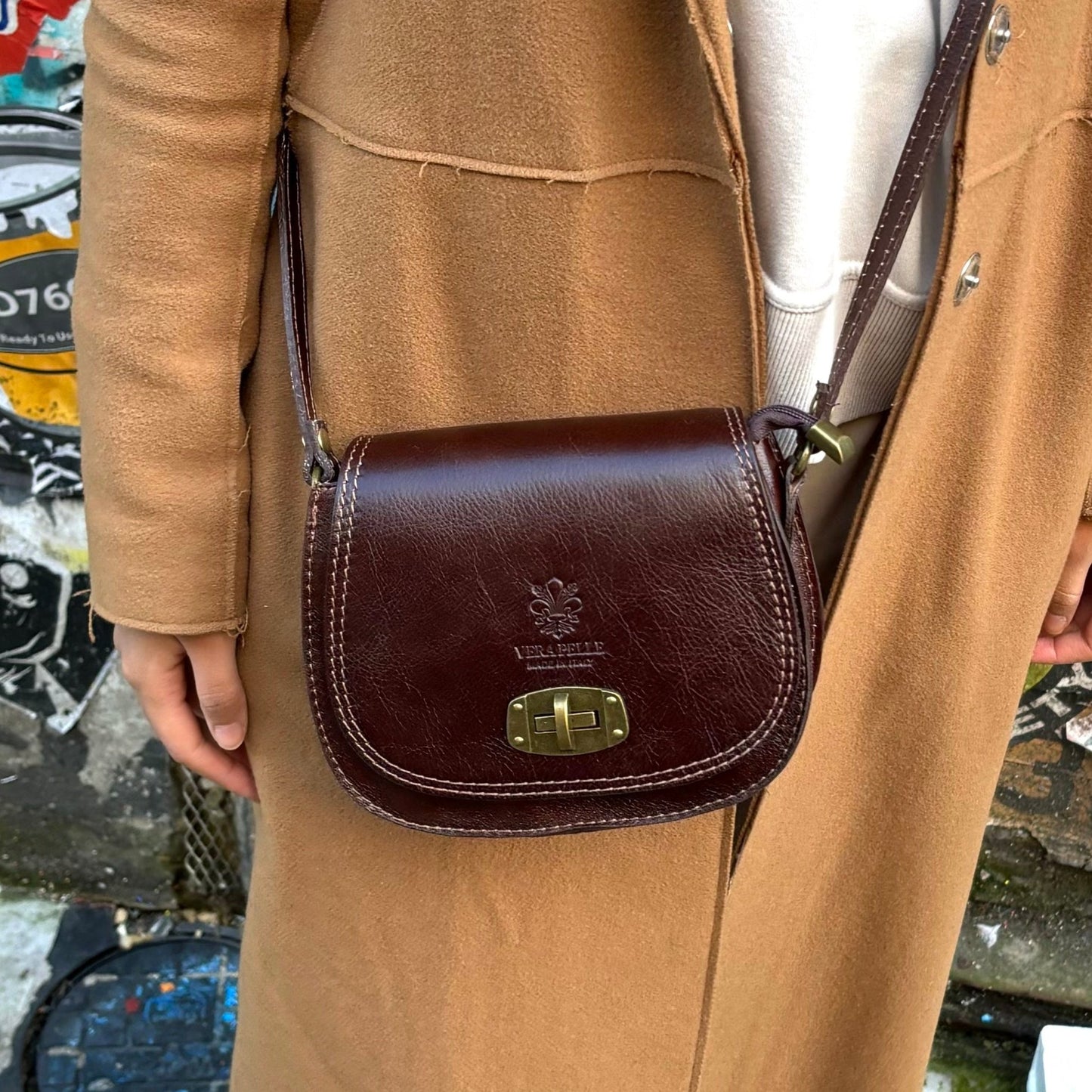 A model wearing a dark tan Cora Vintage Small Leather Crossbody Bag paired with a beige coat, styled for a casual outdoor look.
