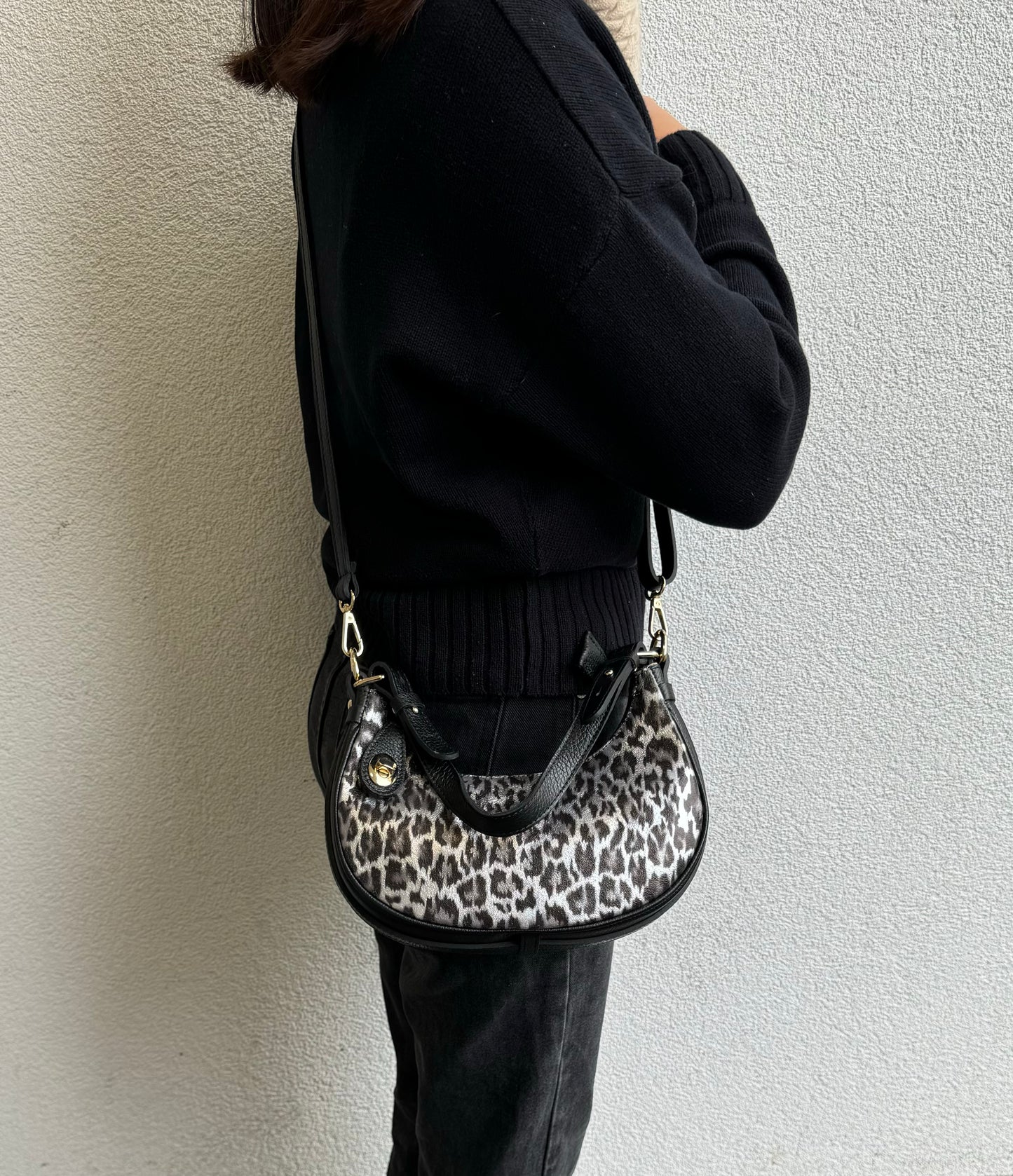 Person wearing a Silver Leopard small half moon shoulder/crossbody bag, side view, with leopard print, black leather accents, and gold-tone hardware against a light-colored wall.