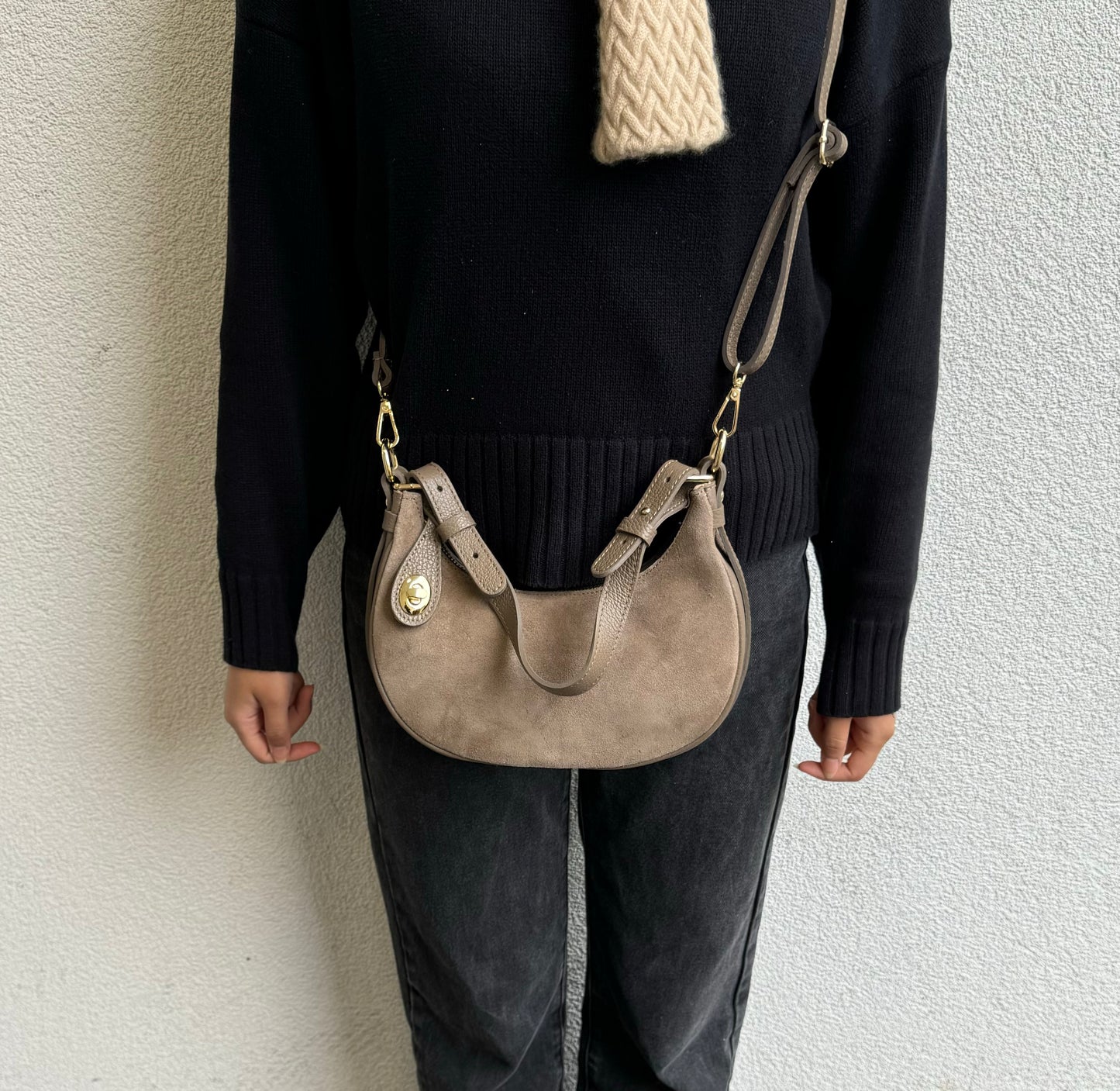 Person wearing a Taupe small half moon shoulder/crossbody bag, front view, with suede texture, gold-tone hardware, and adjustable strap against a light-colored wall.