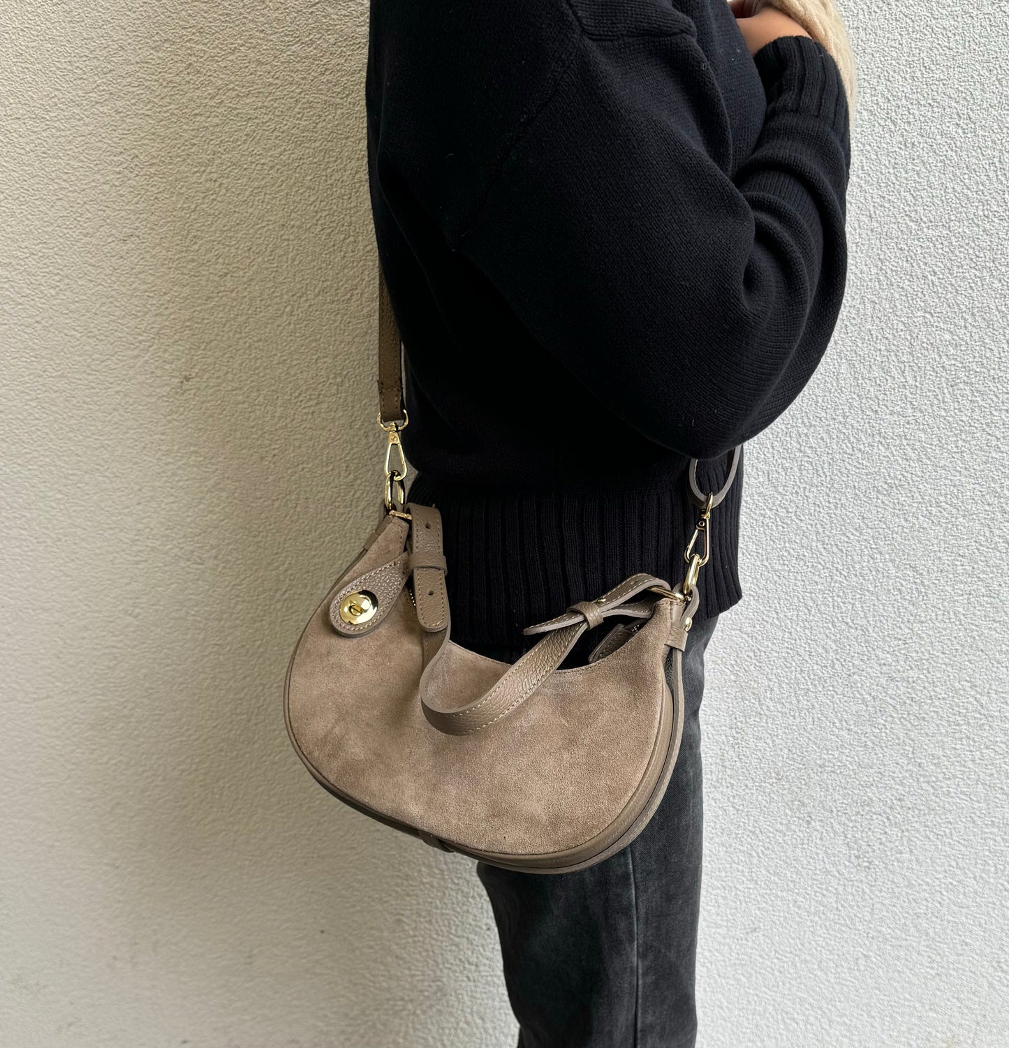 Person wearing a Taupe small half moon shoulder/crossbody bag, side view, featuring suede texture, gold-tone hardware, and adjustable strap against a light-colored wall.