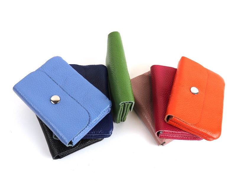 A collection of leather wallets in various colors, including denim blue, navy blue, black, olive green, dusty pink, fuchsia, and bright orange, arranged in a casual, overlapping pattern. Each wallet features a silver snap button.