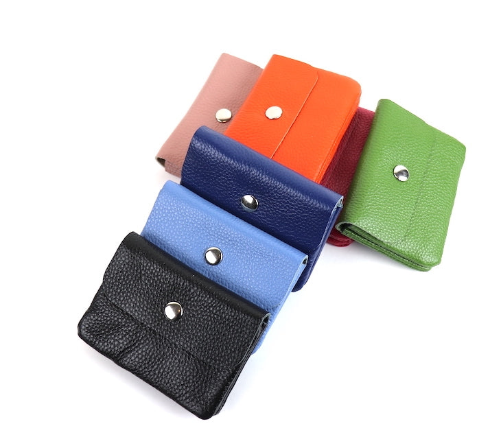 A collection of leather wallets in various colors, including denim blue, navy blue, black, olive green, dusty pink, fuchsia, and bright orange, arranged in a casual, overlapping pattern. Each wallet features a silver snap button.