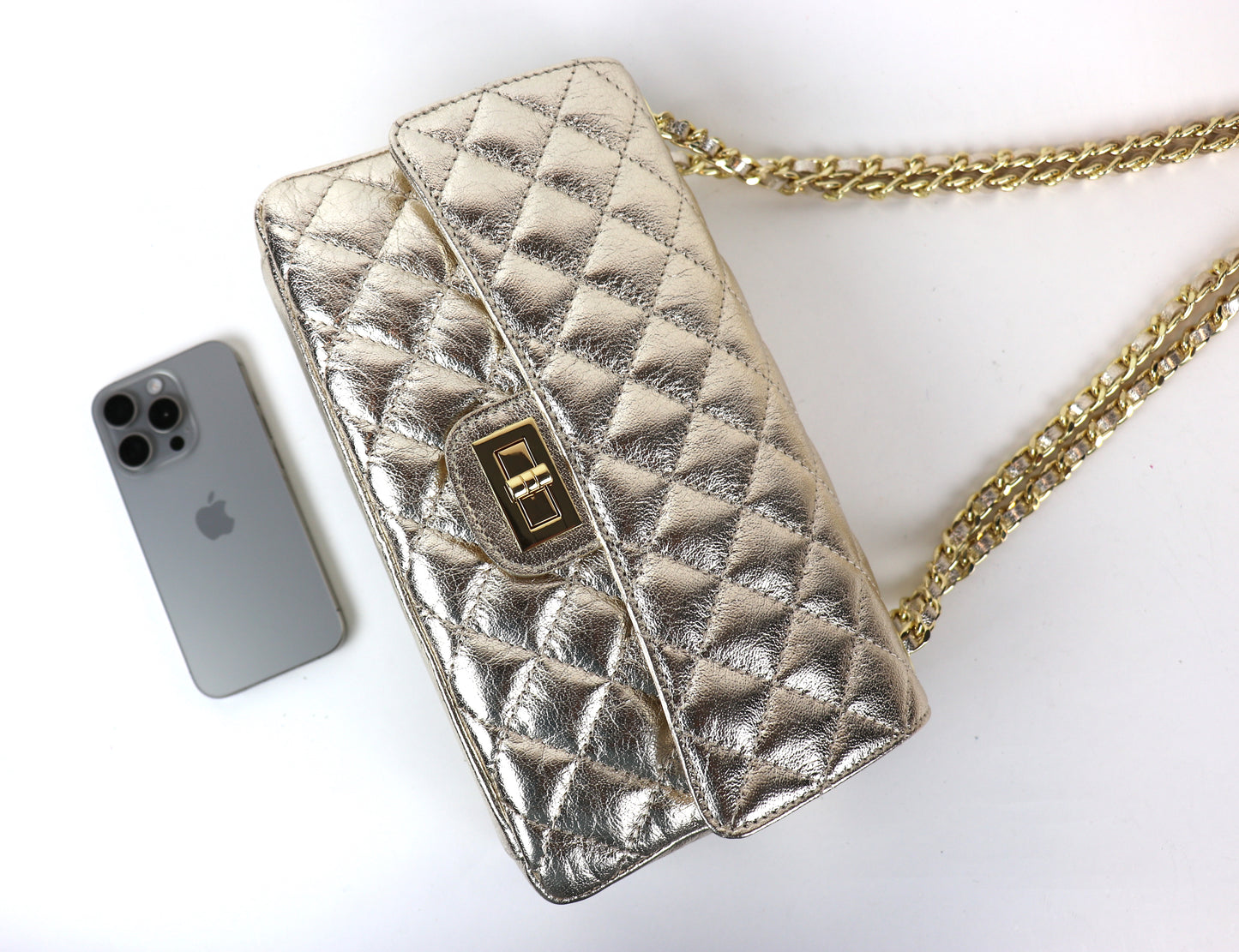 A gold quilted leather bag with a gold twist-lock closure and chain strap, displayed next to a silver iPhone for size reference.






