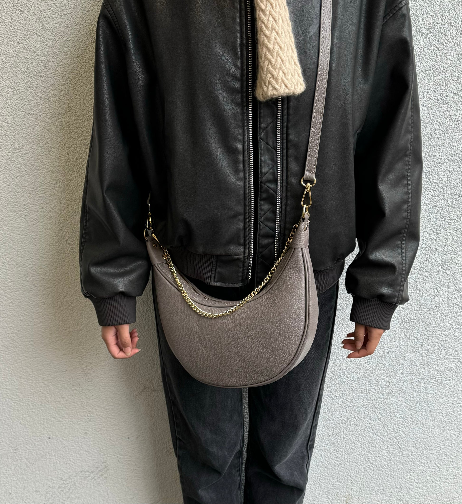 A taupe crescent-shaped leather crossbody bag with a gold chain strap, worn by a person dressed in a black leather jacket. The bag hangs at the model’s side, showcasing its neutral color and structured design, photographed from the front.