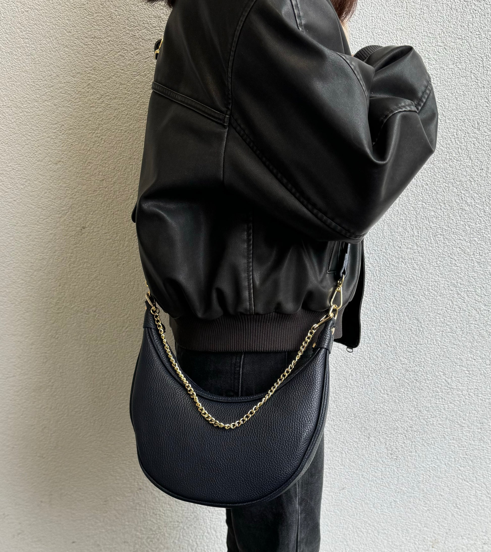 A navy crescent-shaped leather crossbody bag with a gold chain strap, worn by a person in a black leather jacket. The bag hangs at the model’s side, showing the bag’s sleek design and how it complements the casual, stylish outfit.