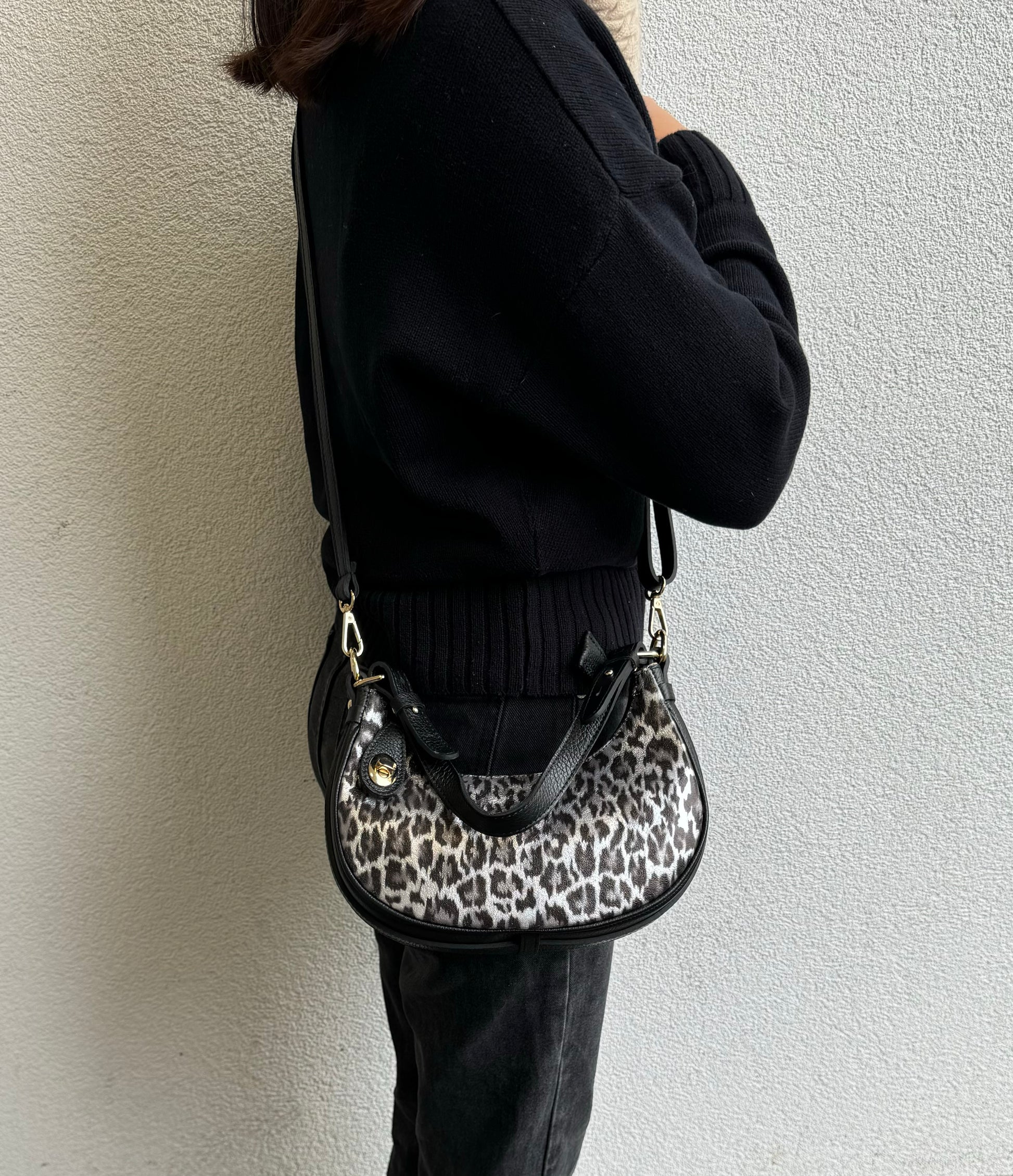 Person wearing a Silver Leopard small half moon shoulder/crossbody bag, side view, with leopard print, black leather accents, and gold-tone hardware against a light-colored wall.

