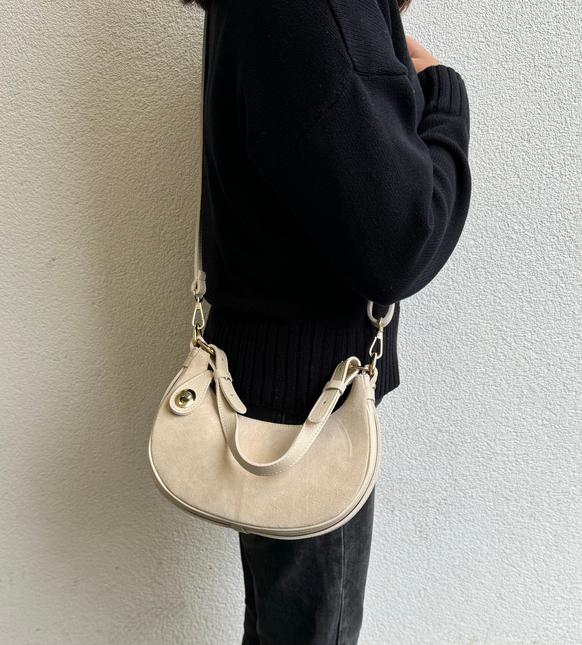 Person wearing a Cream small half moon shoulder/crossbody bag, shown from the side, with a gold-tone clasp and adjustable strap. The bag is slung over the shoulder against a neutral wall background.