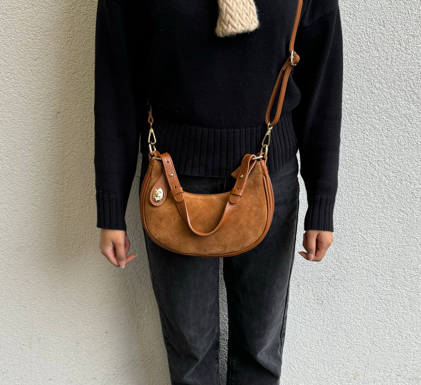 Person wearing a Tan small half moon shoulder/crossbody bag, front view, with suede texture, gold-tone hardware, and adjustable strap against a light-colored wall.

