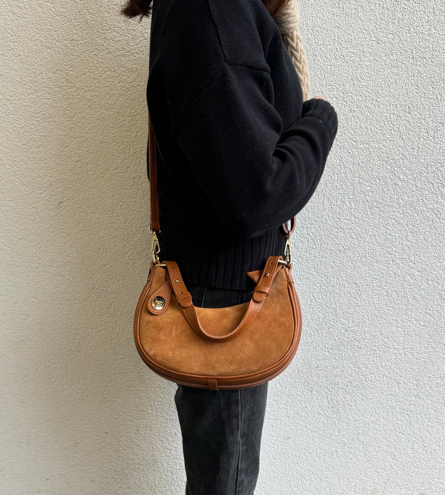 Person wearing a Tan small half moon shoulder/crossbody bag, side view, with suede texture, gold-tone hardware, and adjustable strap against a light-colored wall.