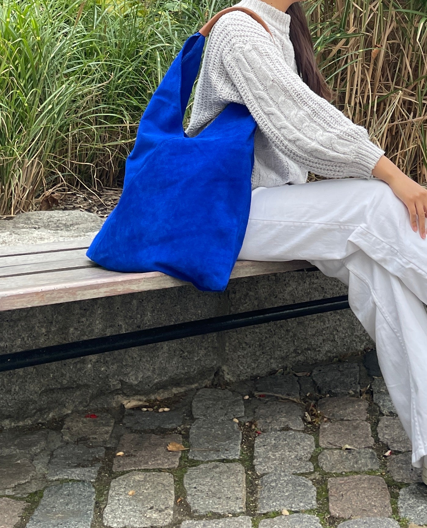 Model sitting on a bench with the Royal Blue Samantha Large Tote Laptop Bag placed behind them, featuring brown leather handles, in an outdoor setting surrounded by greenery.