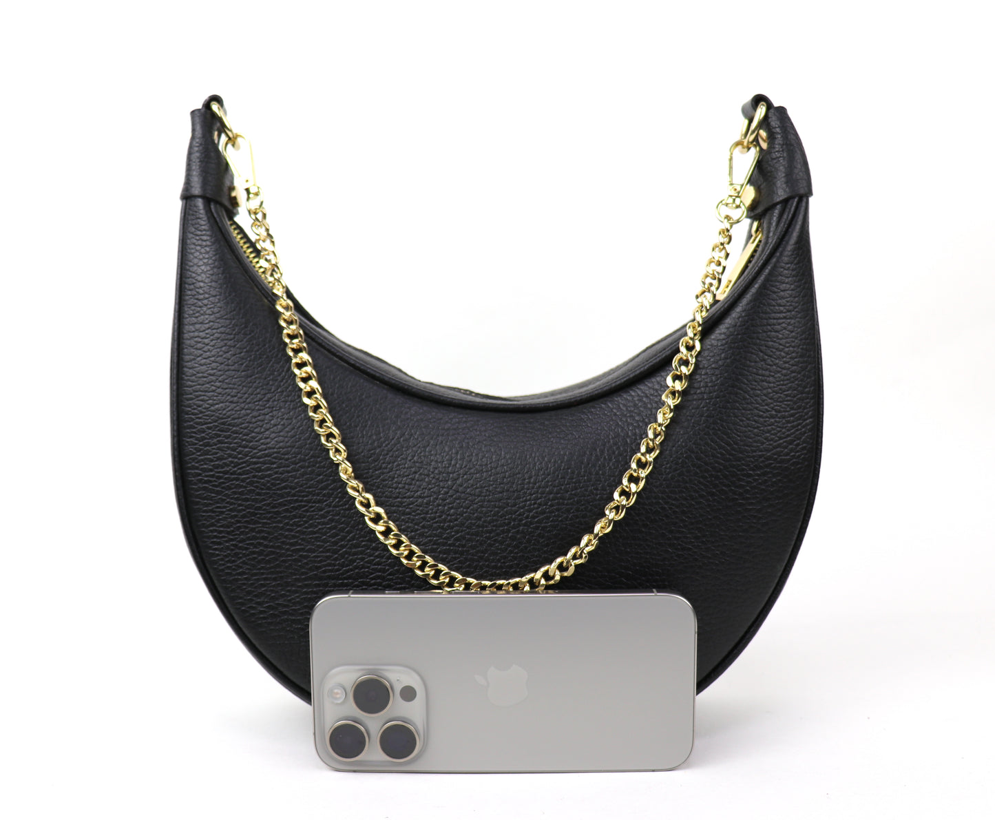 A black crescent-shaped leather crossbody bag with a gold chain strap, displayed against a white background. The texture of the leather is visible, and a silver smartphone is placed in front of the bag for size comparison.