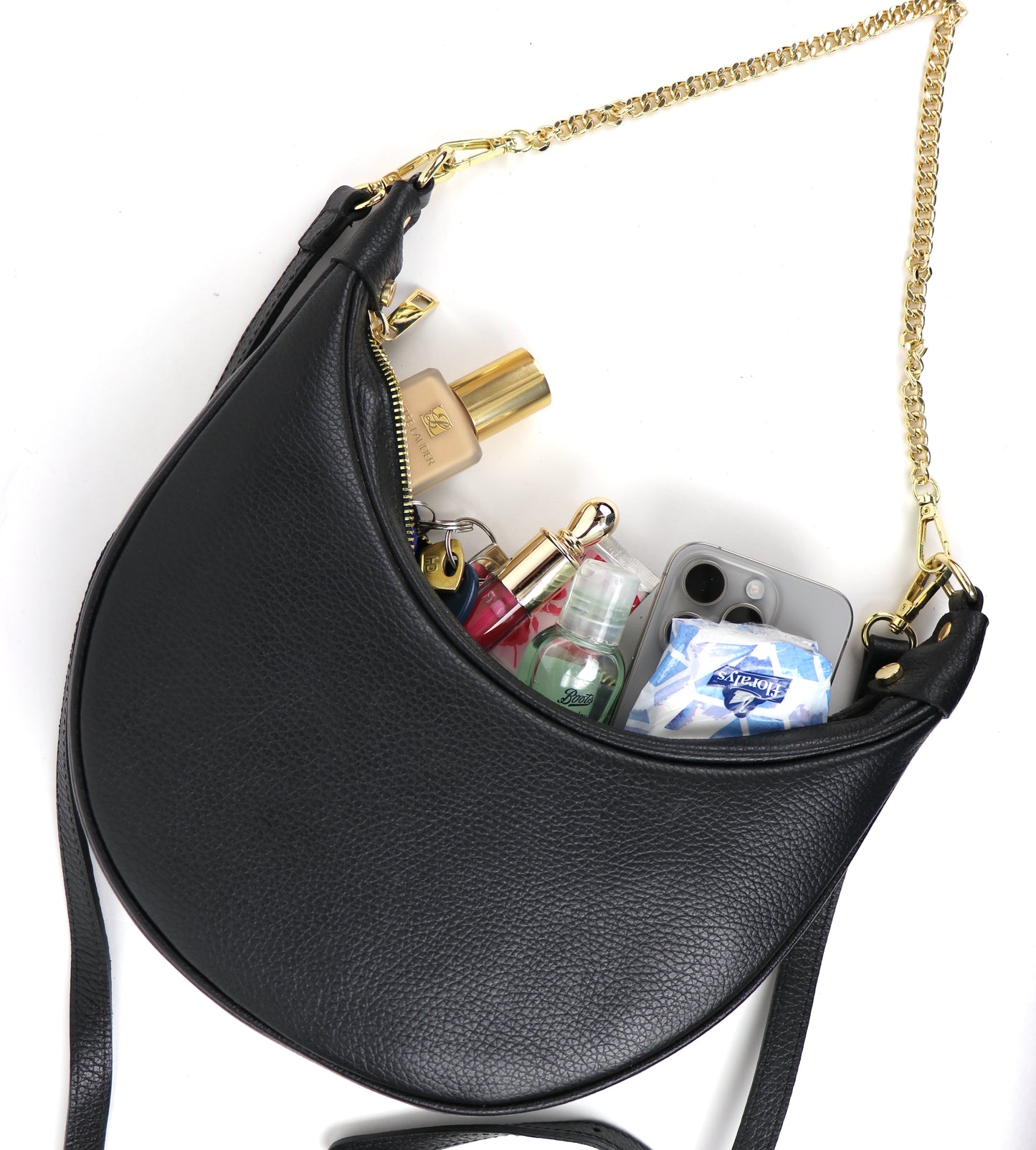 A black crescent-shaped leather crossbody bag with a gold chain strap, photographed from the top angle. The bag is open, revealing several items inside, including a smartphone, perfume bottles, a sanitizer, and a pack of tissues, highlighting the bag's capacity.