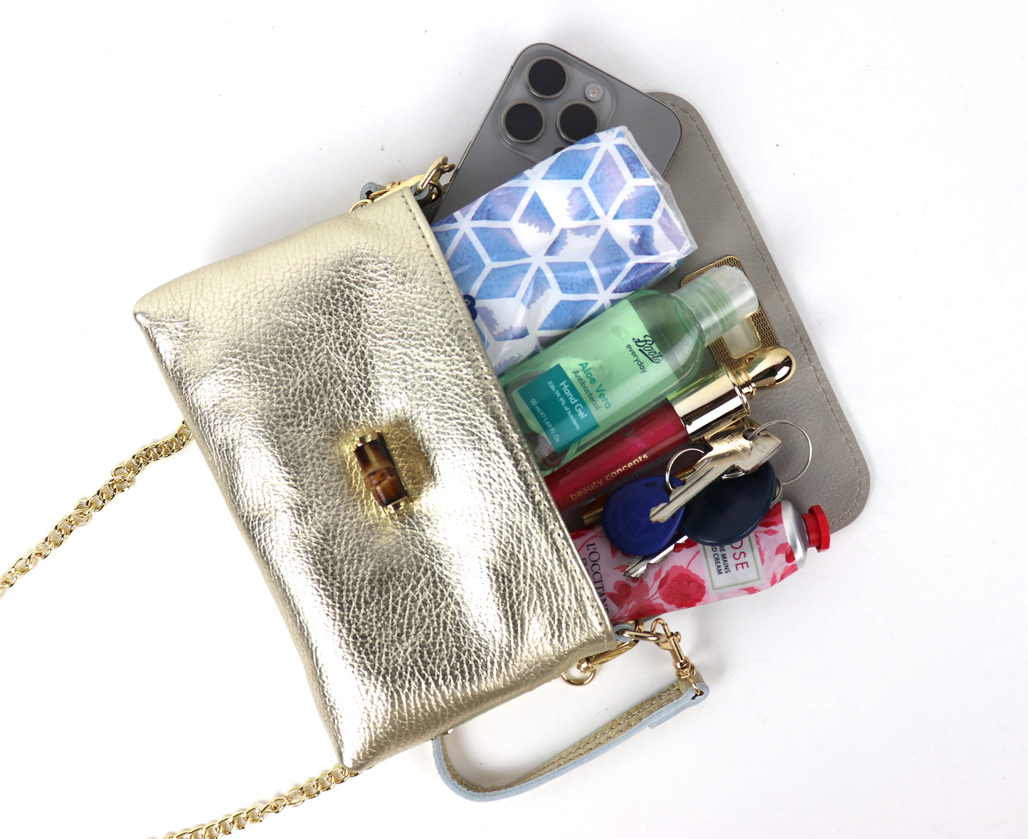 Open gold metallic leather bag with a gold clasp and chain, displaying various items inside, including a smartphone, hand sanitizer, lipstick, keys, and other small essentials. The contents are arranged neatly, showing the capacity and practicality of the bag.

