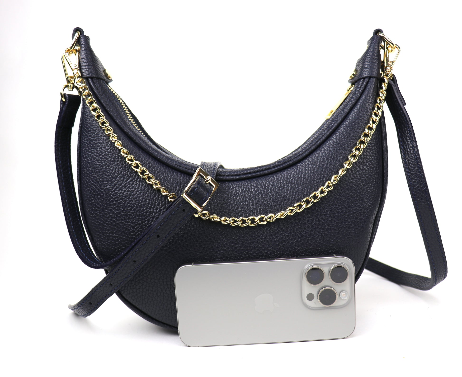 A navy crescent-shaped leather crossbody bag with a gold chain strap, shown from the front. The bag is placed next to a silver smartphone for size reference, showcasing the sleek design and adjustable leather strap.

