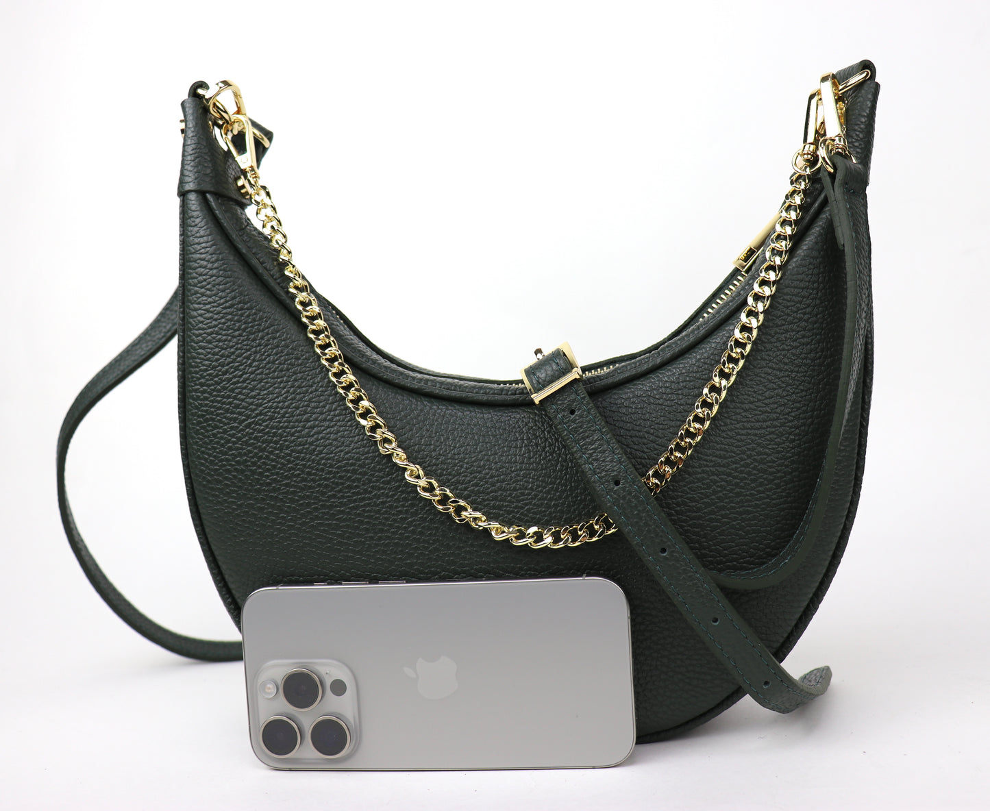 A dark green leather crescent-shaped crossbody bag with a gold chain strap, shown from the front. The bag is accompanied by a silver smartphone placed in front of it for size reference, highlighting the elegant design and deep green color of the bag.