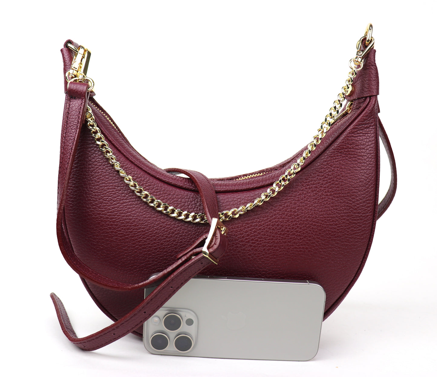 A burgundy leather crescent-shaped crossbody bag with a gold chain strap, photographed from the front. The bag is shown with a silver smartphone placed in front of it for size comparison, highlighting the rich color and elegant design of the bag.